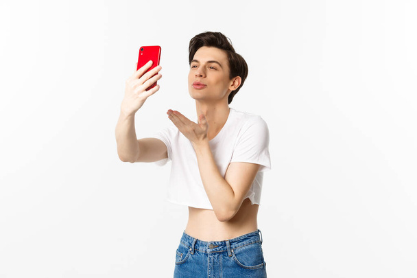piękny gej człowiek w zboże top wysyłanie powietrze pocałunek w telefon aparat biorąc selfie lub wideo czat na smartfon stojąc nad biały tło - Zdjęcie, obraz