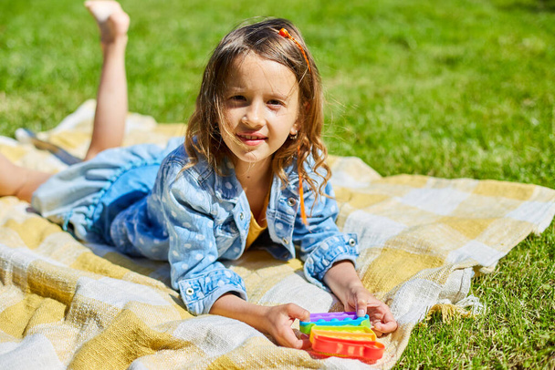 Girl lies on a blanket on grass outdoors and play pop it, kid hands playing with colorful pop It, fidget toy in the backyard of the house on a sunny summer day, summer time vacation. - Photo, image