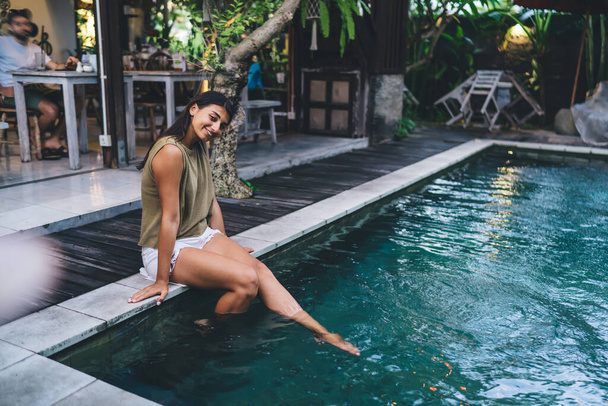 Full length of tanned ethnic woman in t shirt and shorts dipped legs in water and resting by pool with clean water - Foto, Imagen