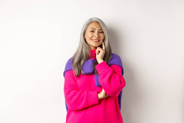 Moda asiática de meia-idade mulher com cabelos grisalhos, sorrindo satisfeito e pensativo, tendo uma ideia, de pé sobre fundo branco - Foto, Imagem