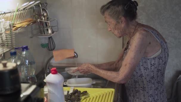 Idosa sênior lavando pratos na cozinha. Triste dona de casa madura limpa os pratos velhos na cozinha. Mulher perturbada com cabelos grisalhos 90 anos lavando utensílios de cozinha em uma lavagem de mãos em casa - Filmagem, Vídeo