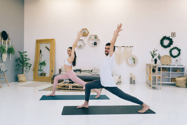 Side view of young flexible couple standing on mats in modern comfort studio and doing lungs with arms raised while looking at camera - Foto, imagen