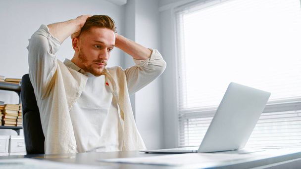 Ο καταθλιπτικός τύπος με το κίτρινο πουκάμισο κοιτάζει το laptop με τη χαμένη σύνδεση στο διαδίκτυο κρατώντας τα χέρια στο κεφάλι - Φωτογραφία, εικόνα