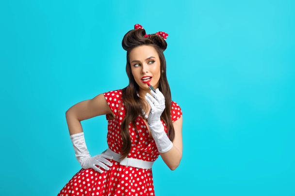Modieuze jonge pinup dame in retro polka dot jurk het aantrekken van make-up, het aanbrengen van lippenstift over blauwe studio achtergrond - Foto, afbeelding
