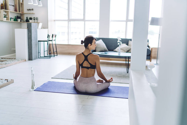 Mindfulned female yogi getting calm inspiration during spiritual meditation in home apartment, back view of woman sitting in lotus pose and thinking about retreat practice for harmony relaxing - Foto, imagen