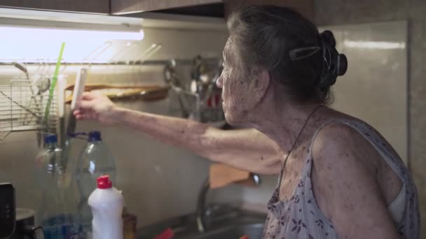 Samotna, smutna starsza kobieta, 90 lat, z siwymi włosami zmuszona zmywać naczynia rękami z powodu ubóstwa w domu, w starej kuchni. Babcia w pracy. Stary brudny dom, kiepskie warunki życia - Materiał filmowy, wideo