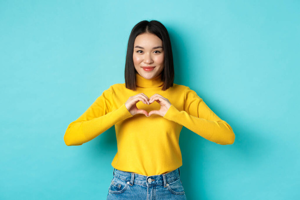 Atrakcyjna japońska dziewczyna w żółtym swetrze, pokazująca gest serca i mówiąca, że cię kocham, patrząca serdecznie w kamerę, stojąca na niebieskim tle - Zdjęcie, obraz