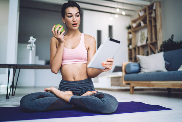 Full body of uzzled sporty slim female in activewear with green apple using tablet and looking at screen perplexingly while sitting in lotus pose on yoga mat at home - Φωτογραφία, εικόνα