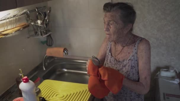灰色の髪の90歳の孤独な悲しい先輩女性は、古いキッチンで自宅で貧困のために彼女の手で料理を洗うことを余儀なくされました。仕事中の祖母。古い汚い家、貧しい生活条件 - 映像、動画