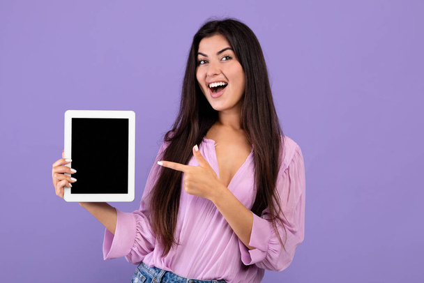 Überprüfen Sie dies. Fröhliche armenische Frau hält und zeigt auf digitale Tablette mit schwarzem leeren Bildschirm, lila Hintergrund - Foto, Bild