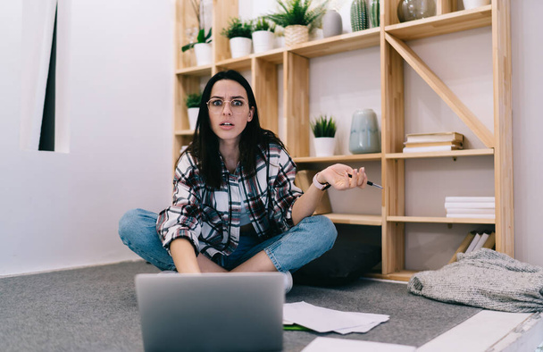 Uncomprehending woman with crossed legs looking at camera with bewilderment while working alone in apartment and surfing computer - Foto, afbeelding