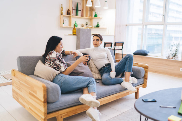 Women sitting on modern sofa with legs crossed and having coffee while spending time together at home in soft daylight - Foto, imagen