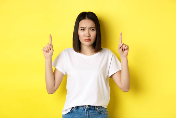 Beauty and fashion concept. Beautiful asian woman in white t-shirt pointing fingers up, standing over yellow background - 写真・画像