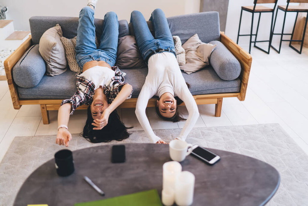 From above of cheerful girlfriends in casual clothes having fun and lying on couch upside down while spending time together - Photo, image