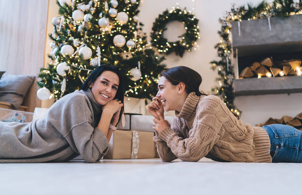 Side view of joyful young female friends in warm clothes lying on floor and having fun after Christmas tree decoration in cozy living room - Фото, зображення