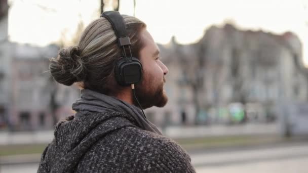 Un uomo barbuto sta ascoltando musica con le cuffie e camminando per la città. Ha i capelli legati. Ritratto di ripresa da un lato. Si sta guardando intorno. 4K - Filmati, video