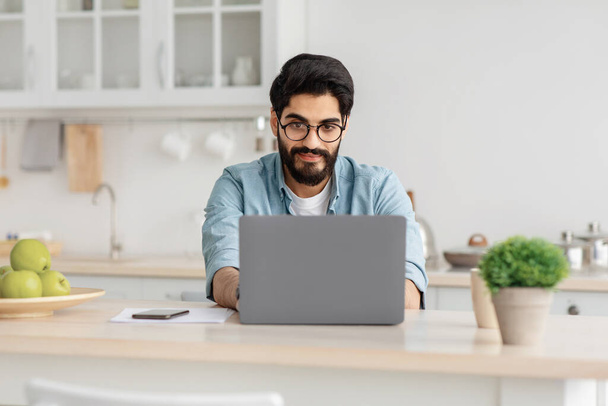 Moderne Fernbedienung. Schöner bärtiger arabischer Mann mit Brille arbeitet am Laptop auf dem Tisch in der Küche, Kopierraum - Foto, Bild