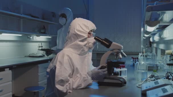 PAN slow de epidemióloga asiática en monos desechables, gafas y mascarilla facial usando microscopio en el laboratorio, luego mirando a la cámara - Metraje, vídeo