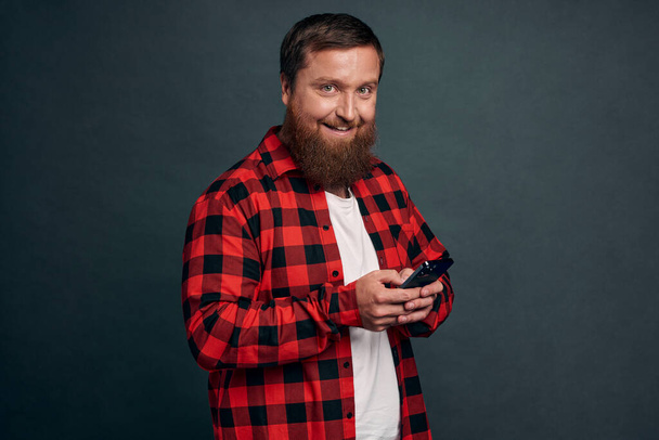 Alegre atractivo sonriente chico feliz con barba en camisa a cuadros rojo, utilizando el teléfono inteligente, mirando cámara entusiasta y alegre, comida para llevar orden con aplicación móvil, de pie fondo gris. - Foto, Imagen