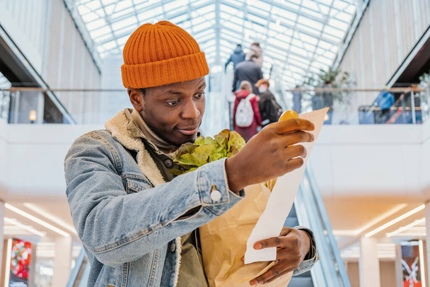 Hombre negro sorprendido mira recibo total con comida en el centro comercial - Foto, Imagen