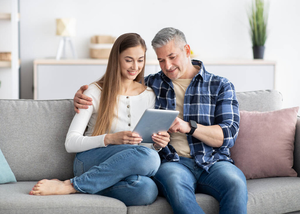 Volwassen echtpaar met tablet zitten op de bank thuis, surfen op internet, chatten online, winkelen in webwinkel - Foto, afbeelding