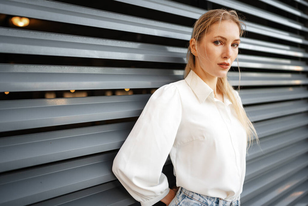 Stylish girl in jeans and white cotton blouse posing near gray wall - Foto, afbeelding