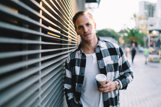 Half length portrait of pensive caucasian man 20s in trendy casual wear holding coffee cup standing on urban setting background, serious male hipster resting during weekend free time on street - Photo, Image