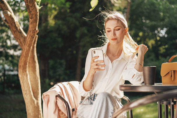 Успішна молода бізнес-леді сидить у відкритому кафе і використовує смартфон
 - Фото, зображення