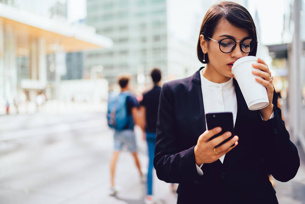 Pensive ethnic businesswoman in formal wear and eyeglasses drinking cup of takeaway coffee while standing on street and browsing smartphone - Foto, afbeelding