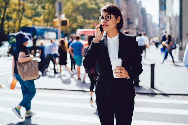 Modern confident female in formal clothes and eyeglasses standing on street and talking on smartphone while drinking hot takeaway coffee - Photo, Image