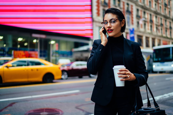 Elegant business lady communicating on cellphone while standing on pavement near busy road holding cup of coffee and looking at camera - Φωτογραφία, εικόνα