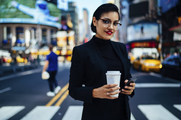 Joven emprendedora sonriente con elegante atuendo y anteojos sosteniendo el teléfono celular y la taza de café mientras está parada en el paso de peatones y mirando la cámara en la ciudad de Nueva York - Foto, Imagen