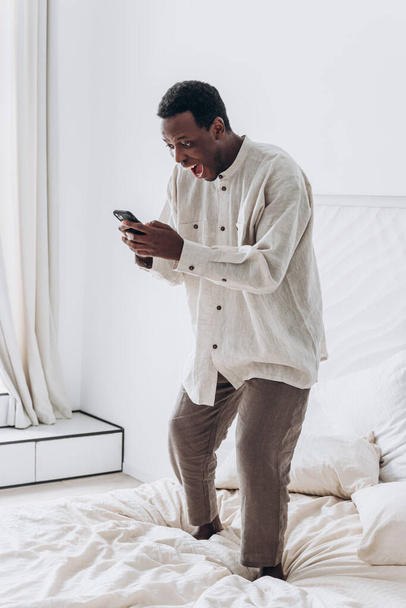 drôle afro-américain gars saute avec téléphone sur grand lit - Photo, image