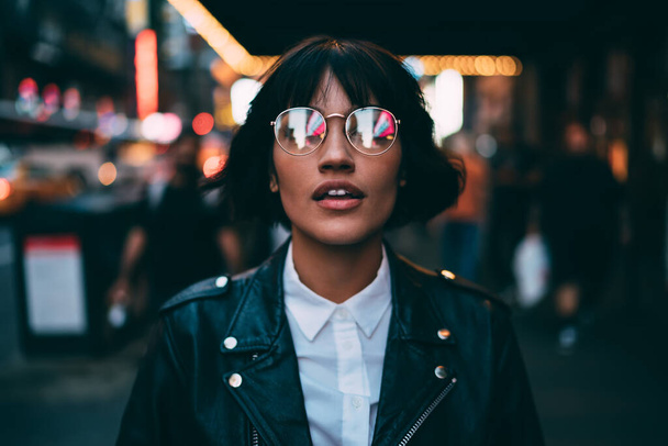 Caucasian woman in optical spectacles with neon reflection of lights standing at urbanity during travel vacations for visiting New York, attractive hipster girl in trendy apparel going out - Photo, image