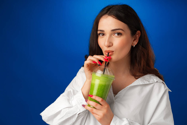 Young beautiful woman in white shirt drinking green smoothie against blue background - Foto, Imagen