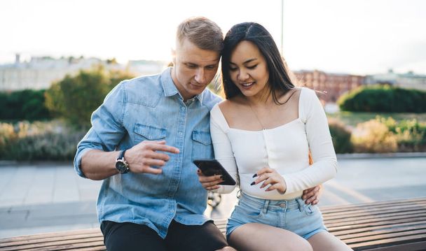 Smiling multiracial couple in casual clothes sitting on wooden bench and browsing smartphone while hugging in sunny day in park - Photo, Image