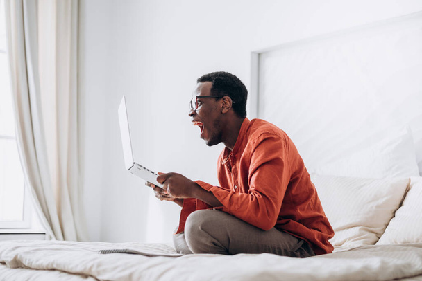 Emotional black man screams holding laptop on large bed - Фото, зображення