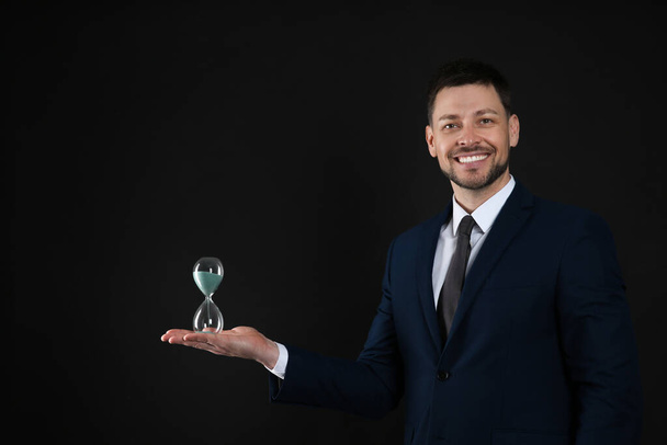 Happy businessman holding hourglass on black background. Time management - Фото, изображение