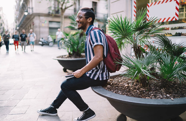 Side view of happy ethnic bearded male in casual clothes and eyewear sitting with backpack and tablet on flowerbed on city street - Foto, immagini