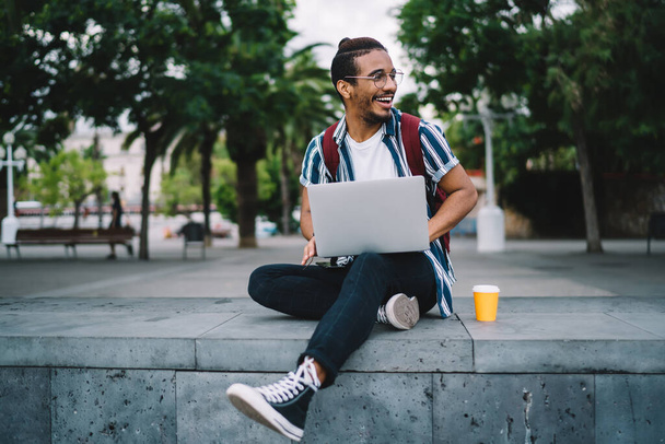 Happy African American male freelancer in casual wear and glasses with backpack using laptop on city street and looking away  while sitting on step with legs crossed and enjoying takeaway coffee on summer day - Foto, Imagem
