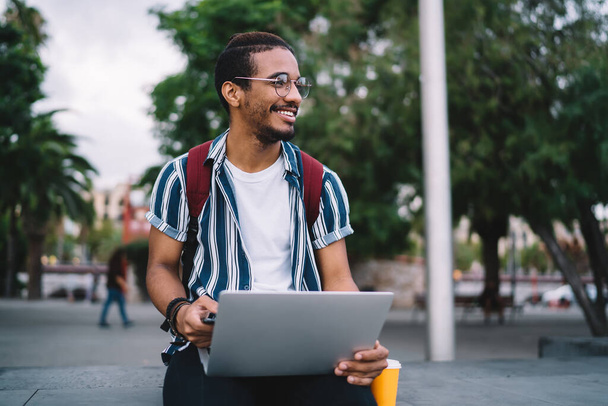 Pozitív etnikai férfi alkalmi nyári viselet és szemüveg dolgozik laptop távolról az utcán, miközben élvezi a kávé elvitel napközben - Fotó, kép