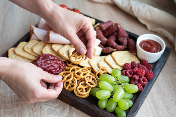 Cocina de fiesta, manos femeninas que ponen el mini pretzel en el tablero de charcutería con salchichas, frutas, galletas y queso, primer plano. - Foto, Imagen