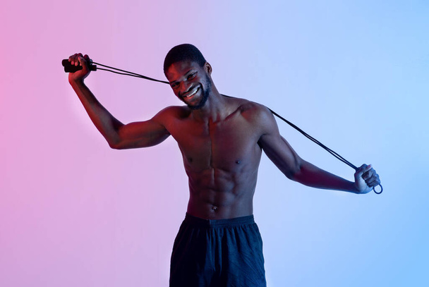 Glücklicher junger schwarzer Sportler mit nacktem Oberkörper posiert mit Springseil in Neonlicht - Foto, Bild