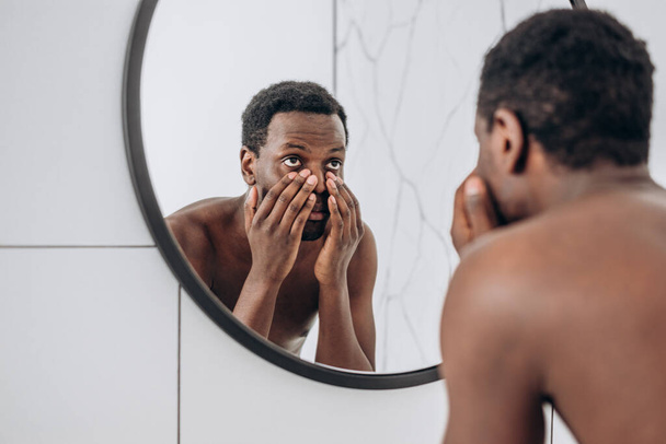 Afroamerikaner blickt im Badezimmer in Spiegel und macht Gesicht - Foto, Bild