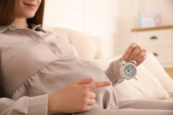 Young pregnant woman pointing at alarm clock near her belly indoors, closeup. Time to give birth - Valokuva, kuva
