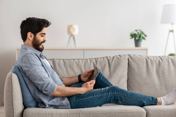 Вид збоку бородатого арабського чоловіка, що сидить на дивані у вітальні, використовуючи цифровий планшет, копіювальний простір
 - Фото, зображення