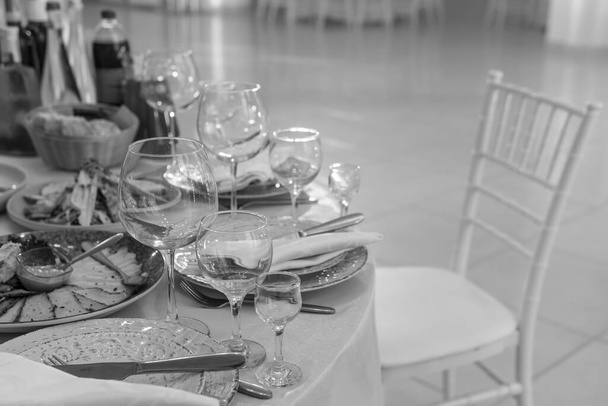 Tisch und Stühle mit Weinglas, Teller voll serviert. Servietten und Besteck. Wenig Licht. Selektiver Fokus. Bereit zum Feiern, zum Feiern. Restaurantdienstleistungen, öffentliche Catering-Unternehmen. Schwarz-Weiß. - Foto, Bild