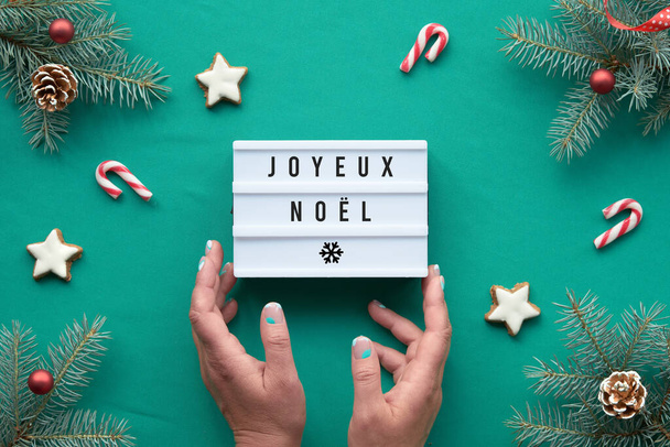 Light box with text Joyeux Noel - Merry Christmas in French language. Hands hold the box. Xmas background, top view on fir twigs with red decorations on vibrant turquoise textile tablecloth. - Photo, Image