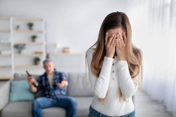 Mature woman crying, husband dinking alcohol, blaming and accusing her at home, free space. Alcoholism and violence - Photo, Image