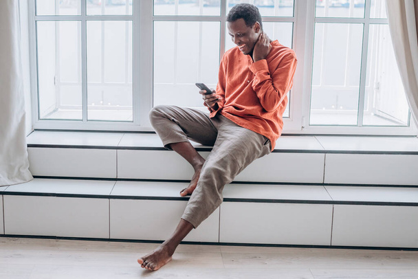 Cheerful black man with smartphone sits on large window sill - Fotografie, Obrázek
